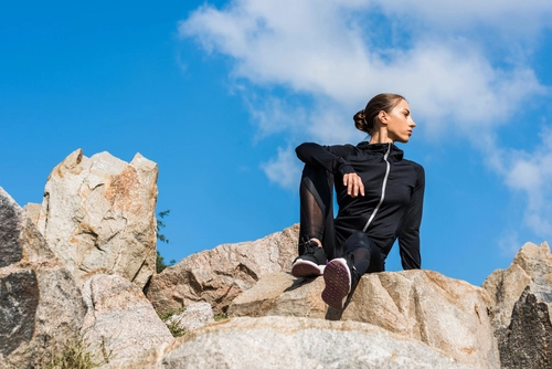 Eine Frau in schwarzem Sportoutfit sitzt auf einem Felsvorsprung und schaut in die Ferne